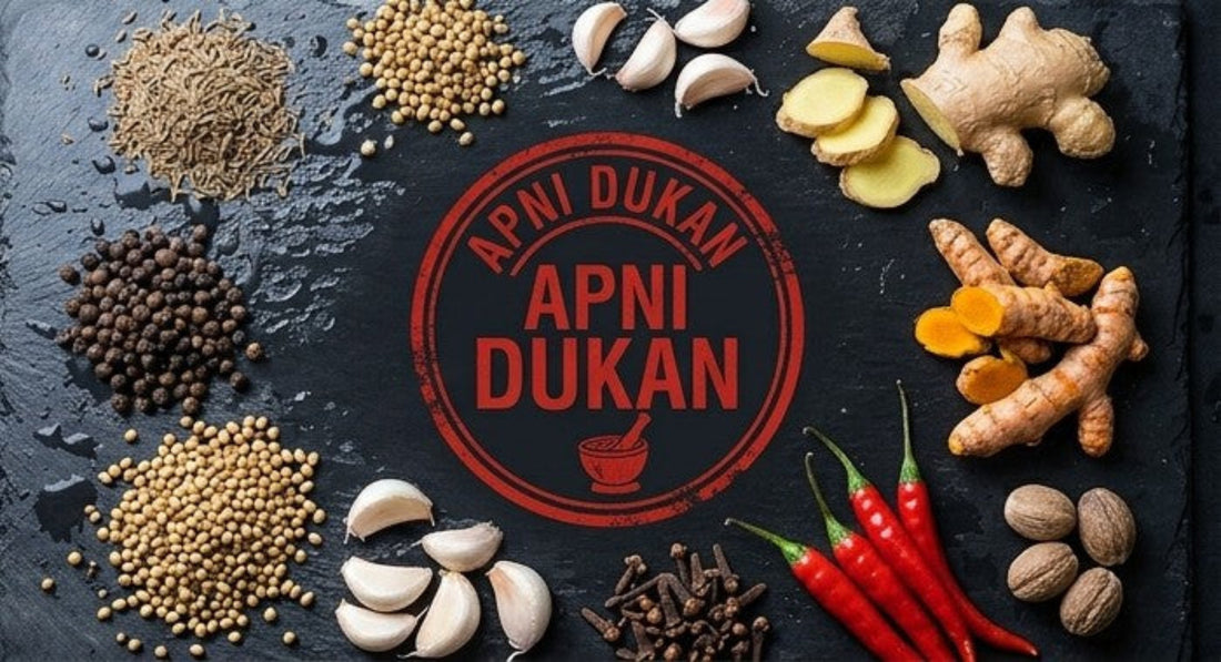 A top-down flat lay of whole spices and ingredients arranged around a central red circular logo reading 'APNI DUKAN'. Visible ingredients include ginger, turmeric, garlic cloves, red chilies, coriander seeds, cumin seeds, black peppercorns, cloves, and nuts on a dark textured surface.
