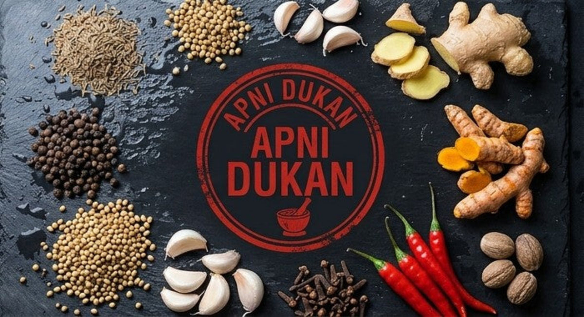 A top-down flat lay of whole spices and ingredients arranged around a central red circular logo reading 'APNI DUKAN'. Visible ingredients include ginger, turmeric, garlic cloves, red chilies, coriander seeds, cumin seeds, black peppercorns, cloves, and nuts on a dark textured surface.